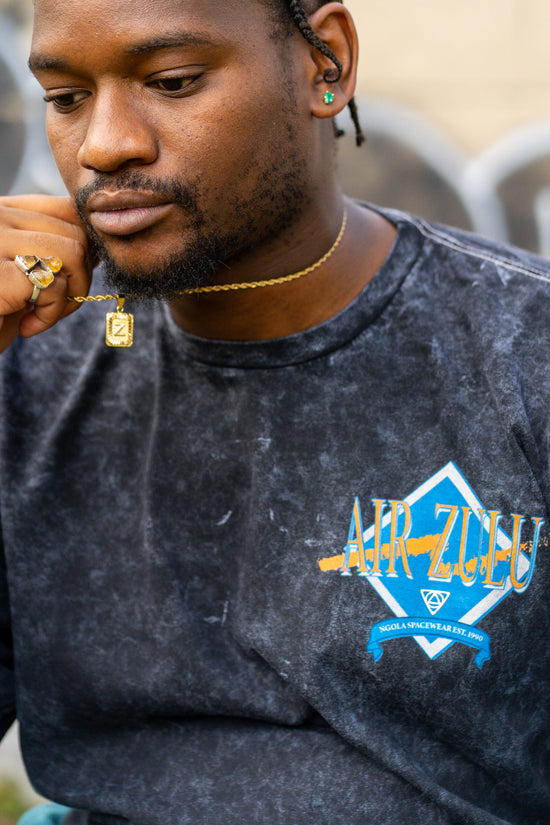 A close-up portrait of a Black man in an Air Zulu shirt, holding a gold necklace with a Z charm.
