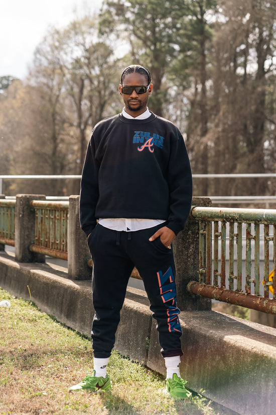 Black man standing in a black colored Air Zulu sweatsuit in front of a railing with trees in the background.
